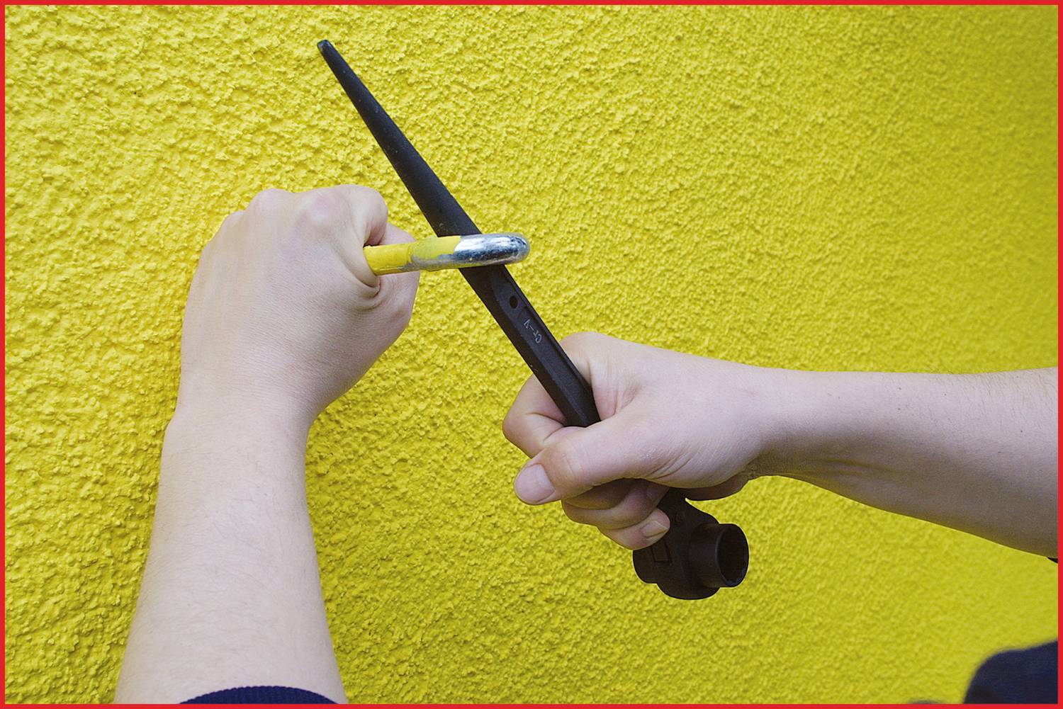 Zwei Hände halten Werkzeuge an gelber Wand, als ob etwas befestigt wird. Eine Hand hält einen Hammer, die andere einen Schraubenschlüssel.