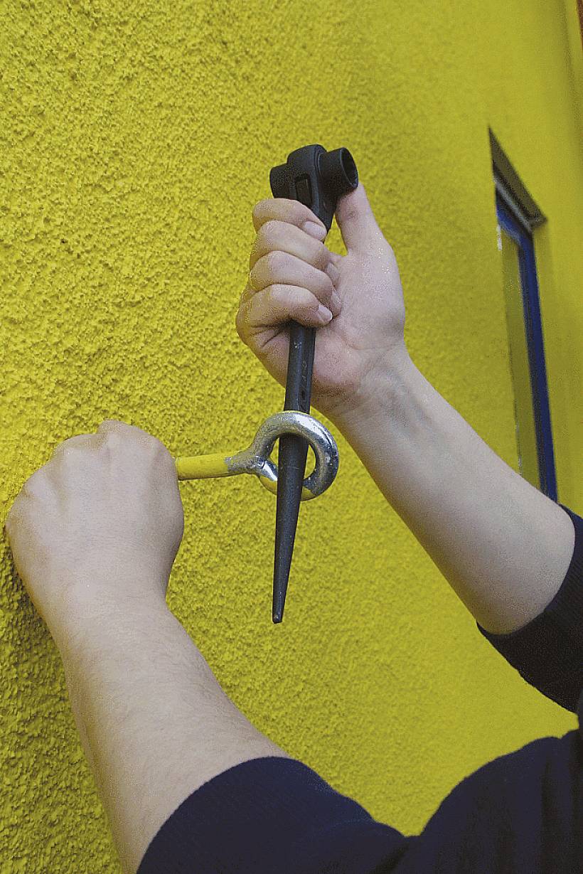 Eine Person befestigt ein Ringankeranker auf einer gelben Wand mit einem Hammer und Ankerstab.