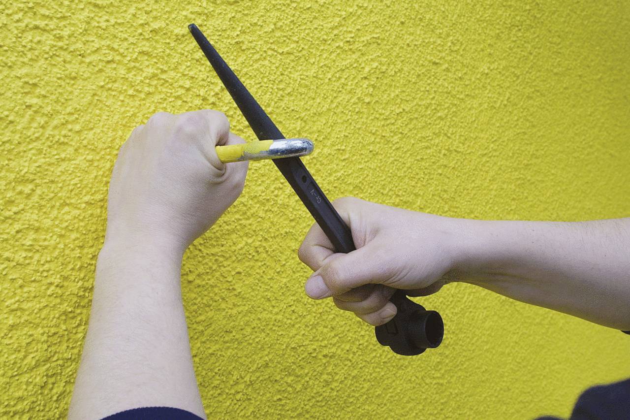 Zwei Hände zeigen, wie man mit einem Hammer und einem Meißel eine gelbe, rauhe Wand bearbeitet.