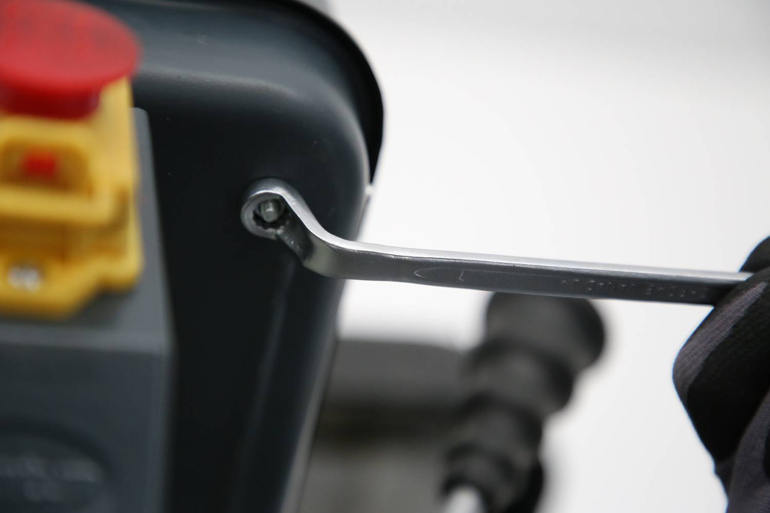 Eine Hand hält einen Schraubenschlüssel, der an einer Schraube eines grauen Geräts arbeitet. Daneben ist ein roter Knopf sichtbar.