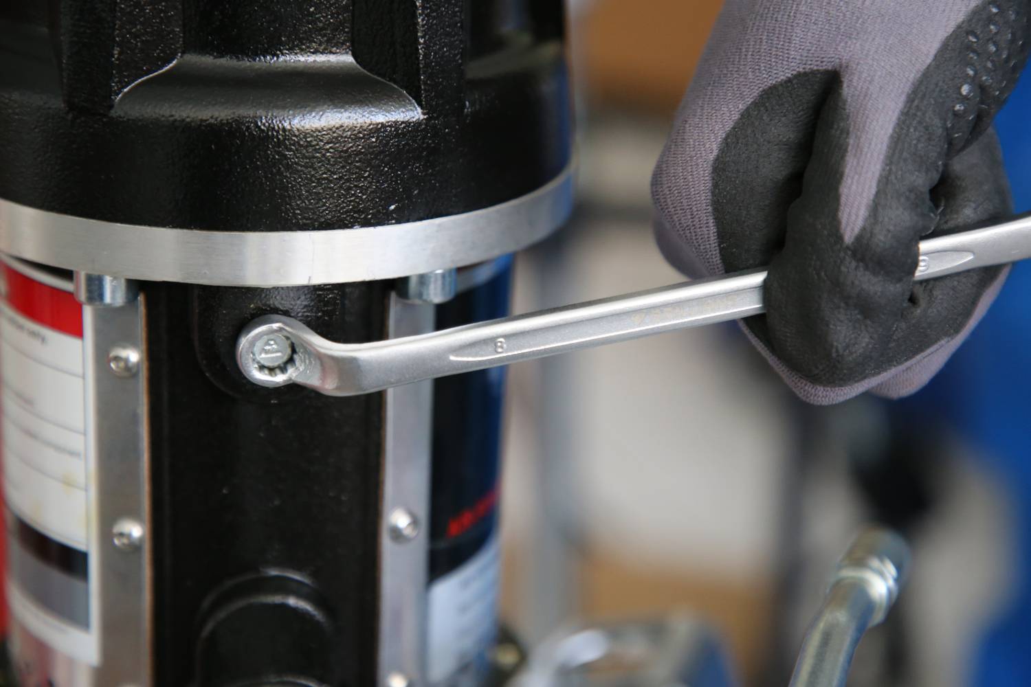 Eine Person zieht mit einem Schraubenschlüssel eine Schraube an einer Maschine fest. Die Hand trägt einen Handschuh.