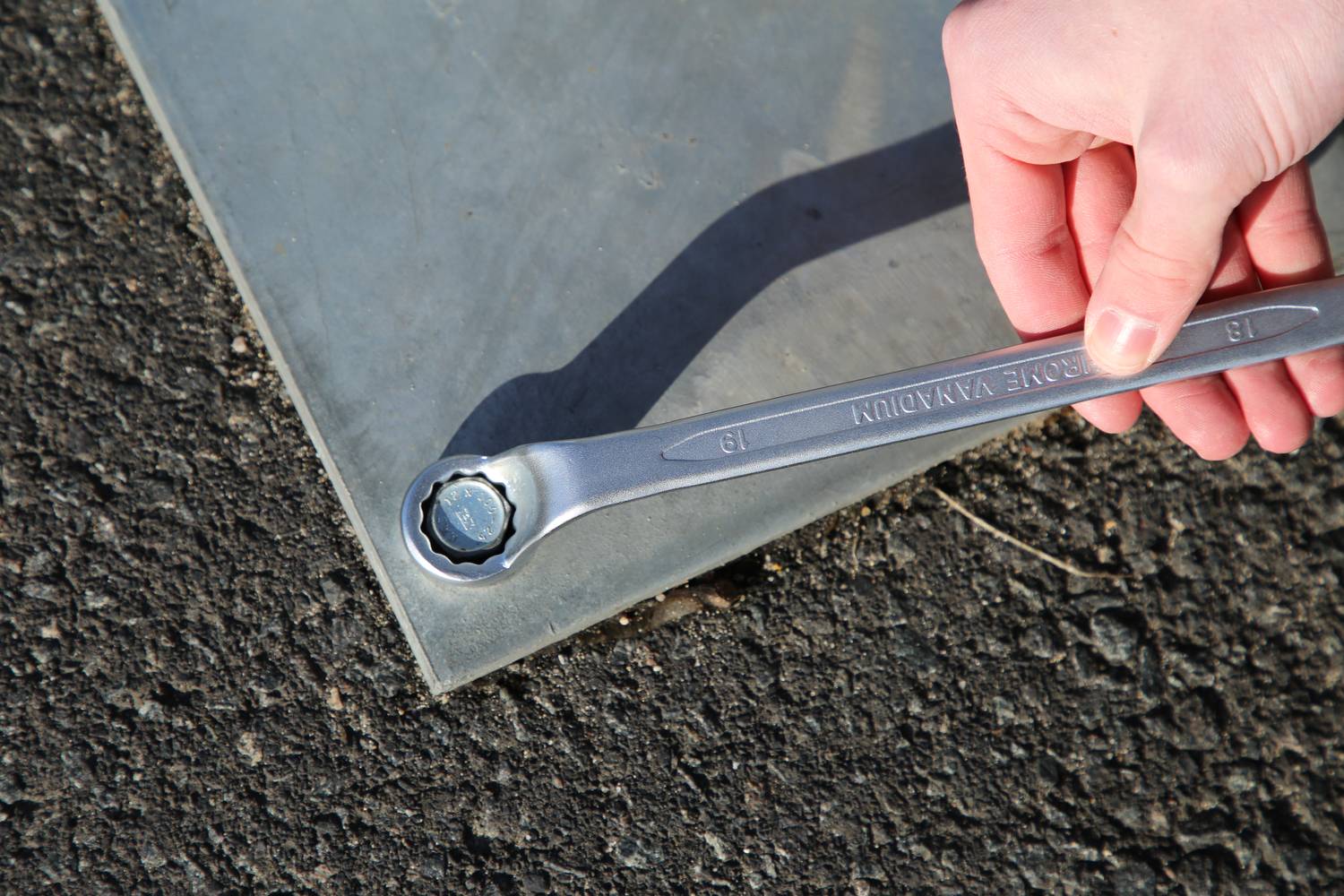 Eine Hand hält einen Schraubenschlüssel, der an einer Schraube auf einer Metallplatte auf Asphalt angelegt ist.