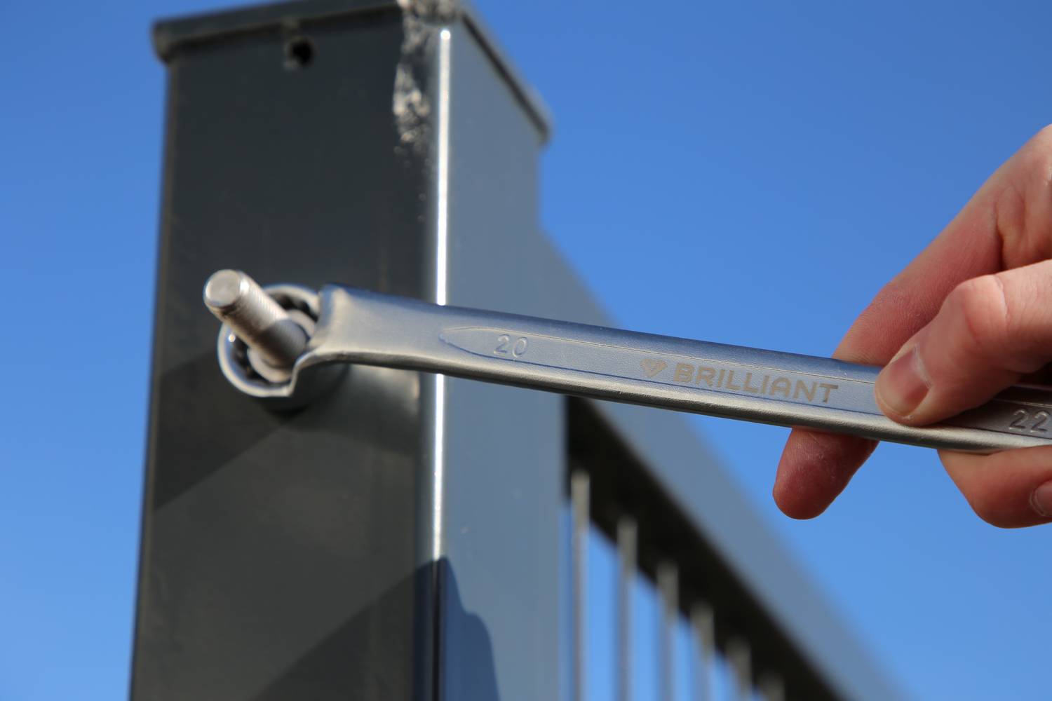 Hand befestigt Schrauben an einem Metallzaunpfosten mit einem Schraubenschlüssel unter blauem Himmel.