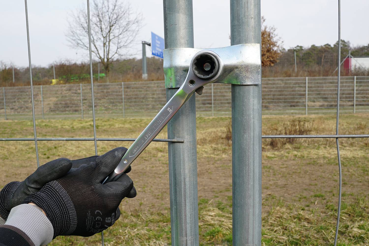 Eine Person zieht mit einem Schraubenschlüssel eine Schraube an, die zwei Metallrohre zusammenhält. Im Hintergrund ist ein Zaun sichtbar.
