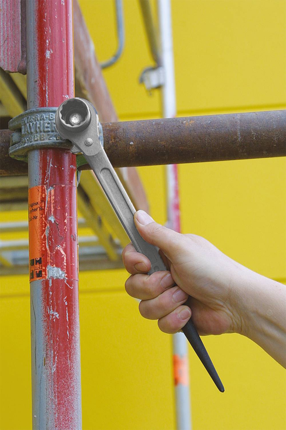 Eine Hand hält einen Schraubenschlüssel und zieht eine Schraube an einem Gerüst an. Der Hintergrund ist leuchtend gelb.