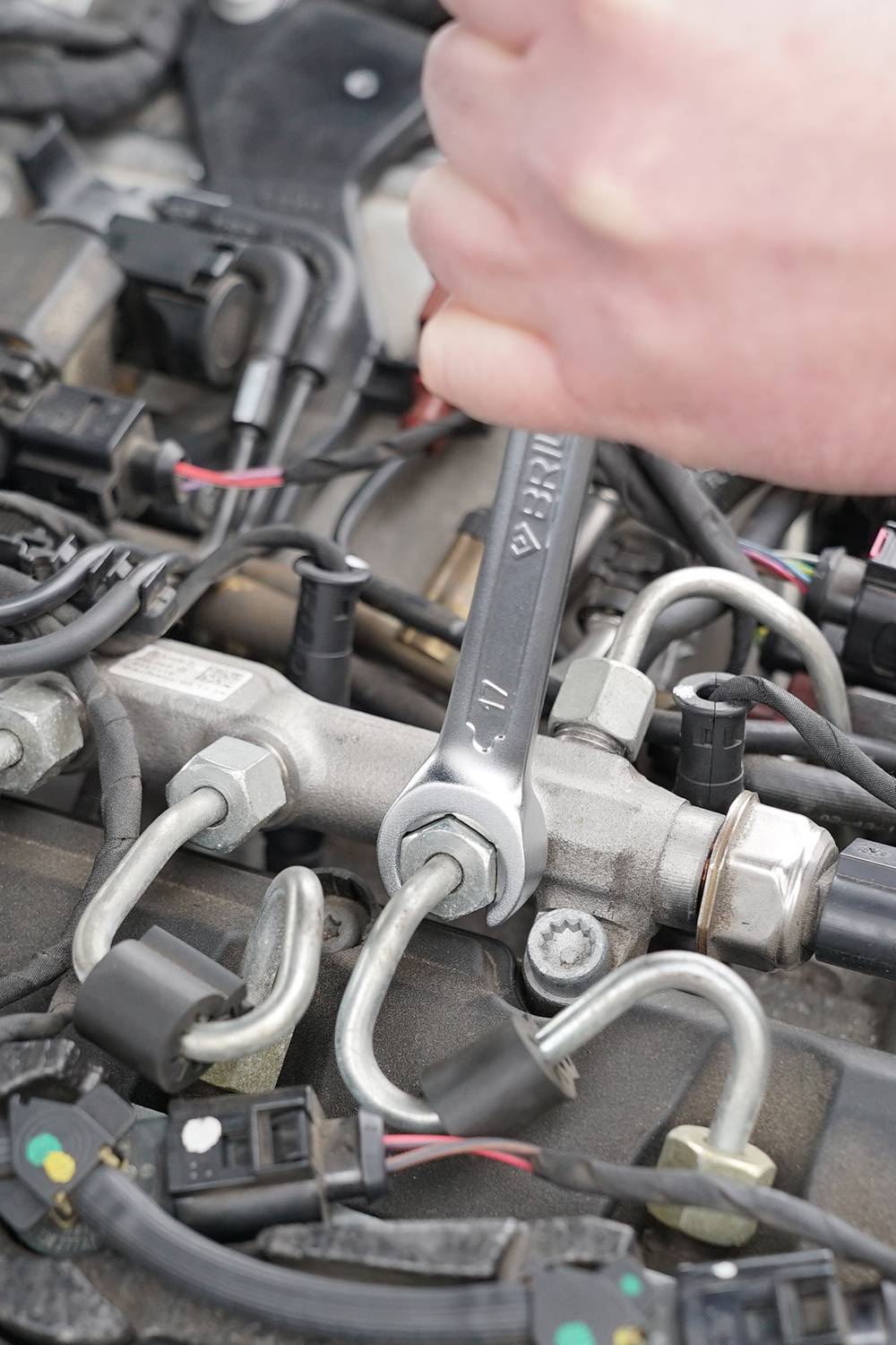 Eine Hand hält einen Schraubenschlüssel und repariert die Einspritzdüsen eines Automotors. Mehrere Kabel und Schläuche sind sichtbar.