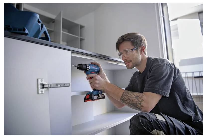 Eine Person verwendet einen elektrischen Bohrer, um einen weißen Schrank zu montieren, trägt Sicherheitsbrille und konzentriert sich darauf, Scharniere im Inneren des Schranks zu befestigen.