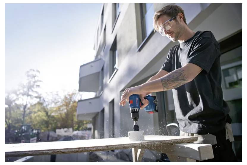 Eine Person mit Sicherheitsbrille benutzt eine Bohrmaschine auf einem Brett außerhalb eines modernen Gebäudes, wobei Sägemehlspray als Schutz sichtbar ist.