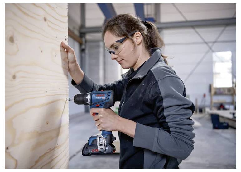 Eine Person mit Sicherheitsbrille verwendet einen kabellosen Bohrer, um an einer Holzplatte in einer Werkstattumgebung zu arbeiten.