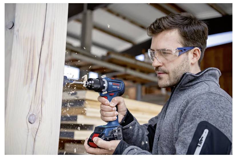 Ein Mann mit Sicherheitsbrille verwendet einen blauen Akkuschrauber, um in einer Werkstattumgebung ein Loch in einen Holzbalken zu bohren.
