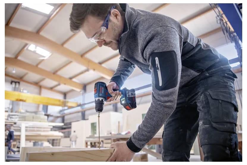 Eine Person benutzt einen Akkuschrauber an Holz in einer Werkstatt, trägt Schutzbrille und eine graue Jacke, umgeben von Baumaterialien.