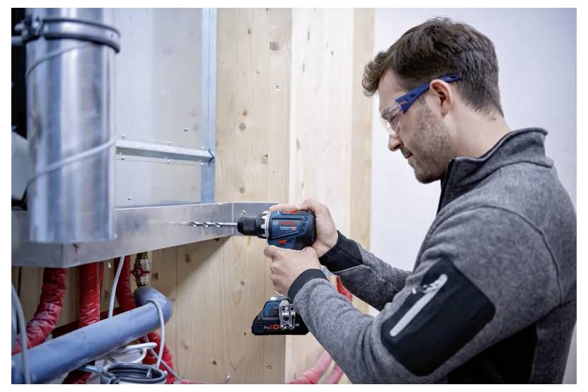 Ein Mann mit Schutzbrille, der eine Bosch-Akku-Bohrmaschine verwendet, um Löcher in einen Metallträger zu bohren, umgeben von freiliegenden Rohren und Holzbalken.