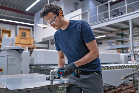 Ein Arbeiter, der eine Schutzbrille und Handschuhe trägt, schneidet ein Metallblech mit einem tragbaren Elektrowerkzeug in einer Industriewerkstatt.