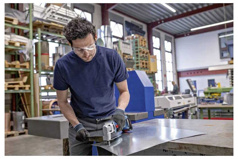 Ein Arbeiter trägt Schutzbrille und Handschuhe und nutzt eine Elektrosäge, um ein Metallblech auf einer Werkbank in einer Werkstatt zu schneiden.