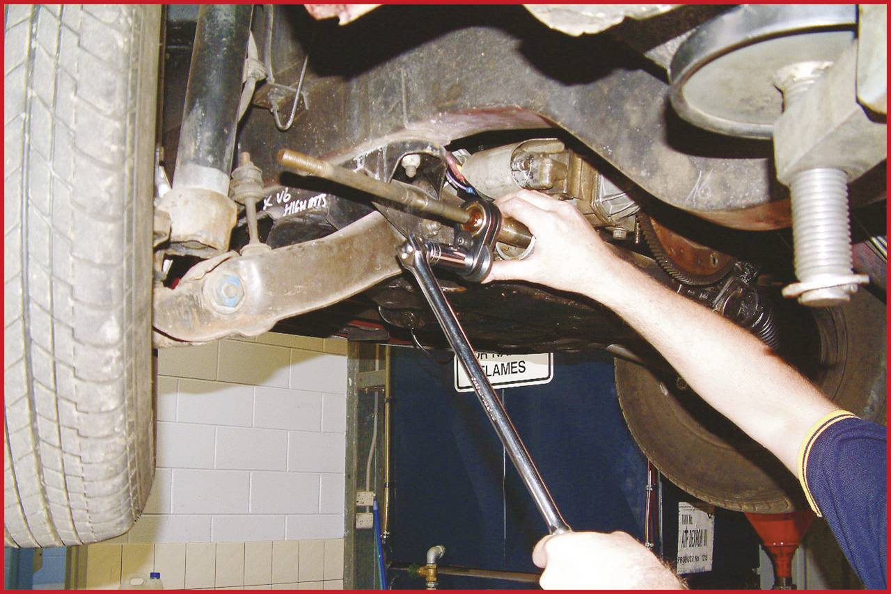 Eine Person repariert unter einem Fahrzeug ein Teil der Aufhängung mit einem Schraubenschlüssel in einer Autowerkstatt.