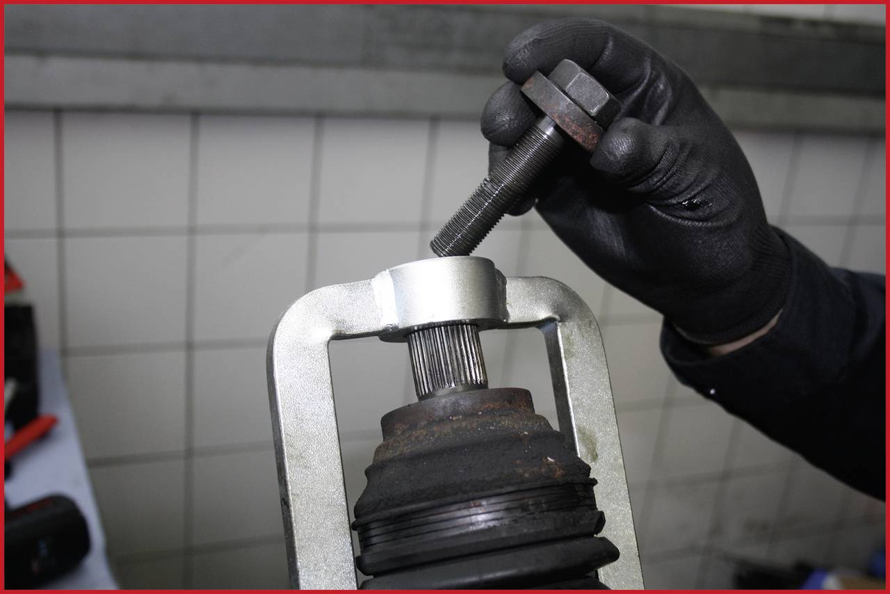 Eine Person mit schwarzen Handschuhen nutzt ein Werkzeug an einem Autoteil in einer Werkstattumgebung.