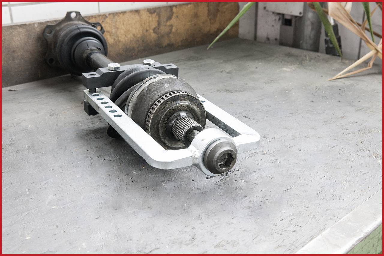 Ein Autoteil liegt auf einem Werkstattboden. Es ist eine Antriebswelle mit sichtbaren Gelenken und Metallkomponenten.