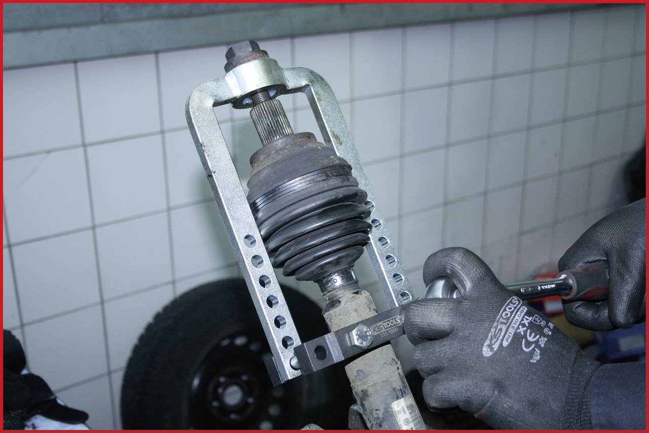 Eine Person demontiert eine Antriebswelle mit einem Abzieher in einer Werkstatt. Im Hintergrund ist ein Reifen zu sehen.