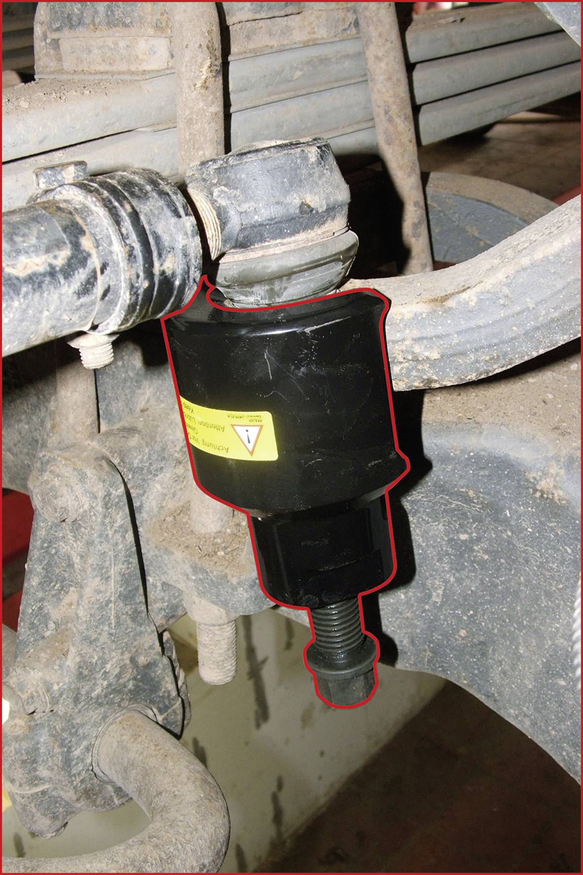 Ein schwarzes Bauteil mit einem gelben Warnetikett ist an einem metallischen Fahrwerksystem einer Maschine oder eines Fahrzeugs befestigt.