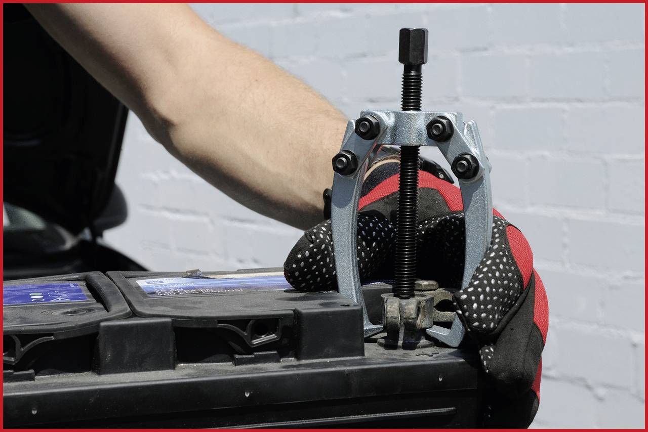 Eine Person mit Handschuhen benutzt einen Abzieher, um eine Batterieklemme von einer Autobatterie zu entfernen.