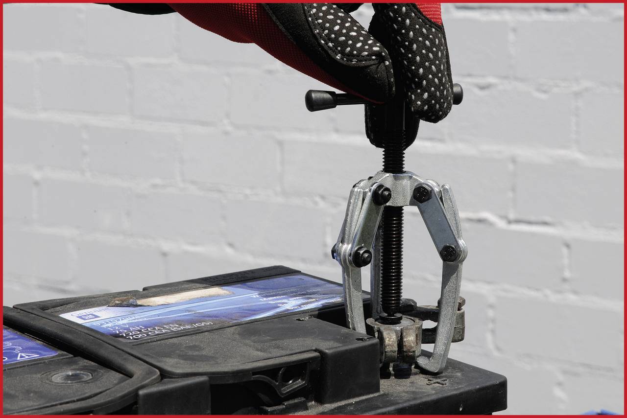 Eine Hand mit Handschuh benutzt einen Abzieher, um einen Polverbinder von einer Autobatterie zu entfernen.