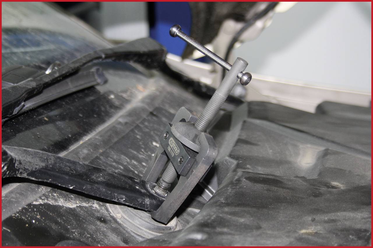 Ein Abzieher-Werkzeug ist an einem Autoteil befestigt, um es zu entfernen. Das Auto und Werkzeug sind staubbedeckt.