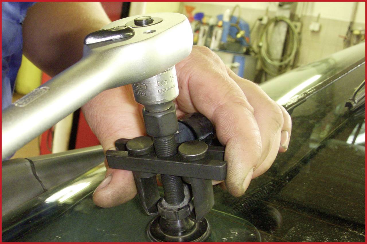 Eine Nahaufnahme zeigt eine Hand, die ein Werkzeug nutzt, um ein Autoteil zu reparieren oder anzupassen. Im Hintergrund sind Werkzeuge sichtbar.