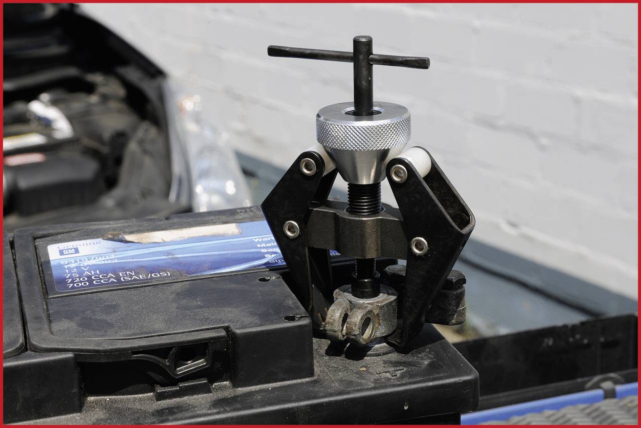 Ein Werkzeug zieht einen Polverbinder von einer Autobatterie ab. Die Batterie liegt in einem geöffneten Motorraum.