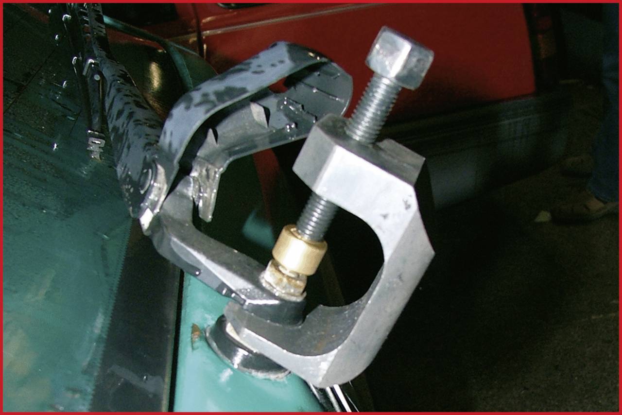 Metallwerkzeug mit Schraubzwinge befestigt an einem grünen Fahrzeugteil vor einem roten Hintergrund.