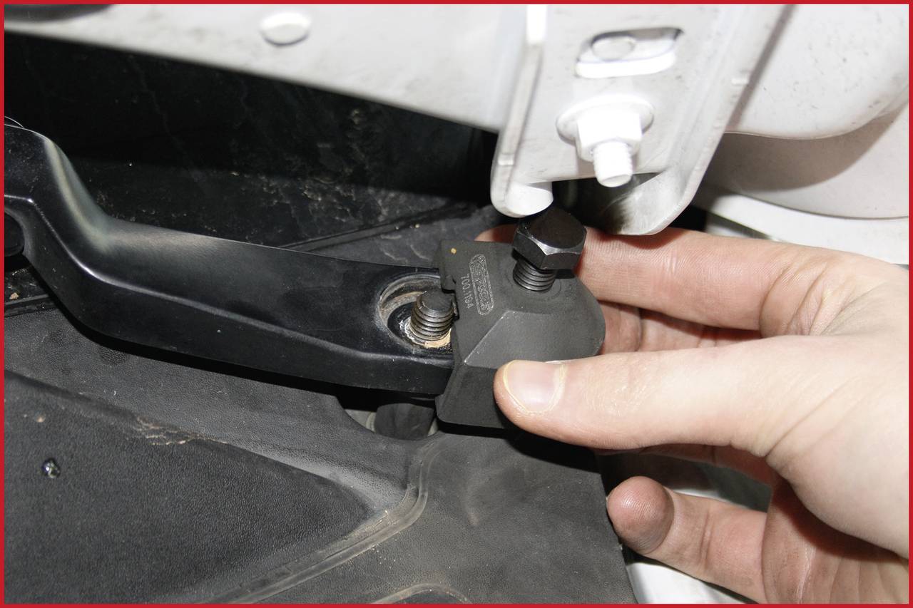 Eine Hand justiert ein Autoteil durch Drehen eines Schraubenschlüssels an einem schwarzen Hebel unter einer Motorhaube.