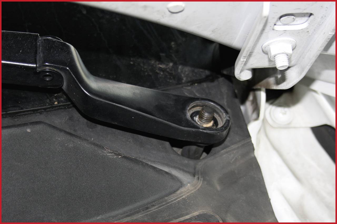 Ein schwarzer Metallarm verbunden mit einer Schraubmutter in einem Fahrzeug, vermutlich Teil der Aufhängung oder Türmechanik.