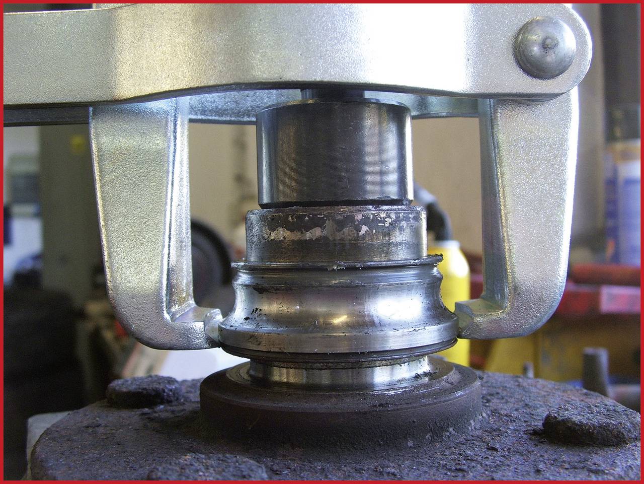Nahaufnahme einer Hydraulikmaschinenkomponente, die Metallteile bearbeitet. Zylinder übt Druck auf Lager aus. Werkstatt im Hintergrund.