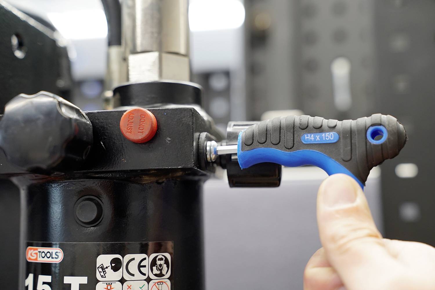 Eine Hand benutzt einen blauen Inbusschlüssel, um eine Schraube an einer schwarzen hydraulischen Presse anzuziehen.