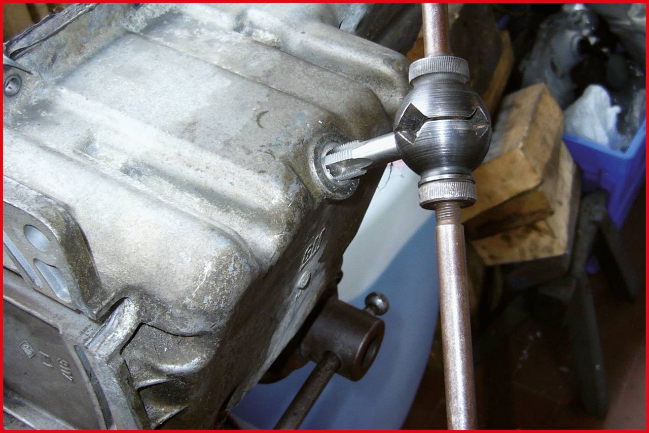 Eine Metallvorrichtung mit einem Gewindebohrer, der in ein Maschinengehäuse eingedreht wird. Im Hintergrund sind Werkzeuge sichtbar.