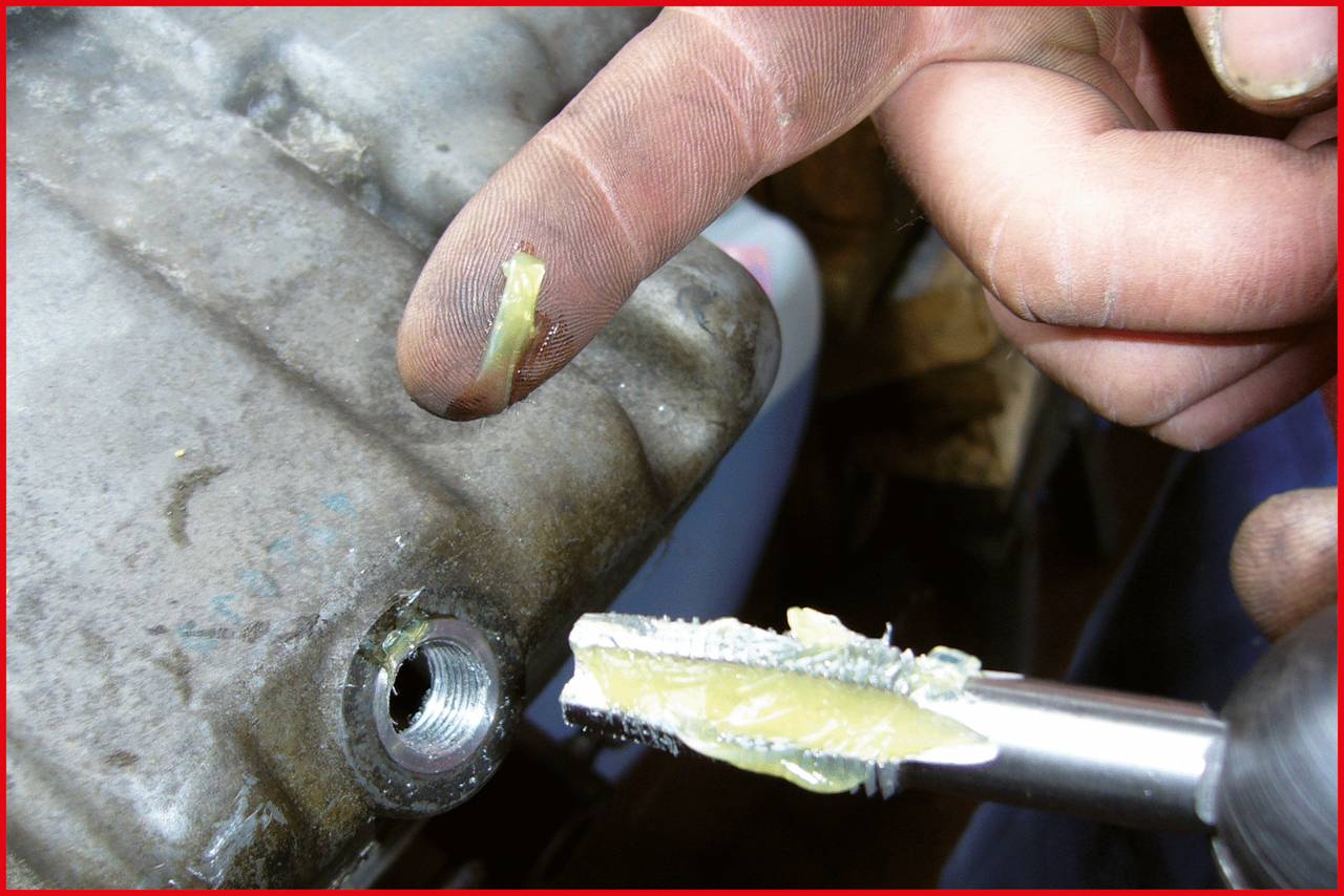 Eine Hand zeigt Fettreste an einem Bohrer und einem Metallteil. Die Szene deutet auf Wartungs- oder Reparaturarbeiten hin.