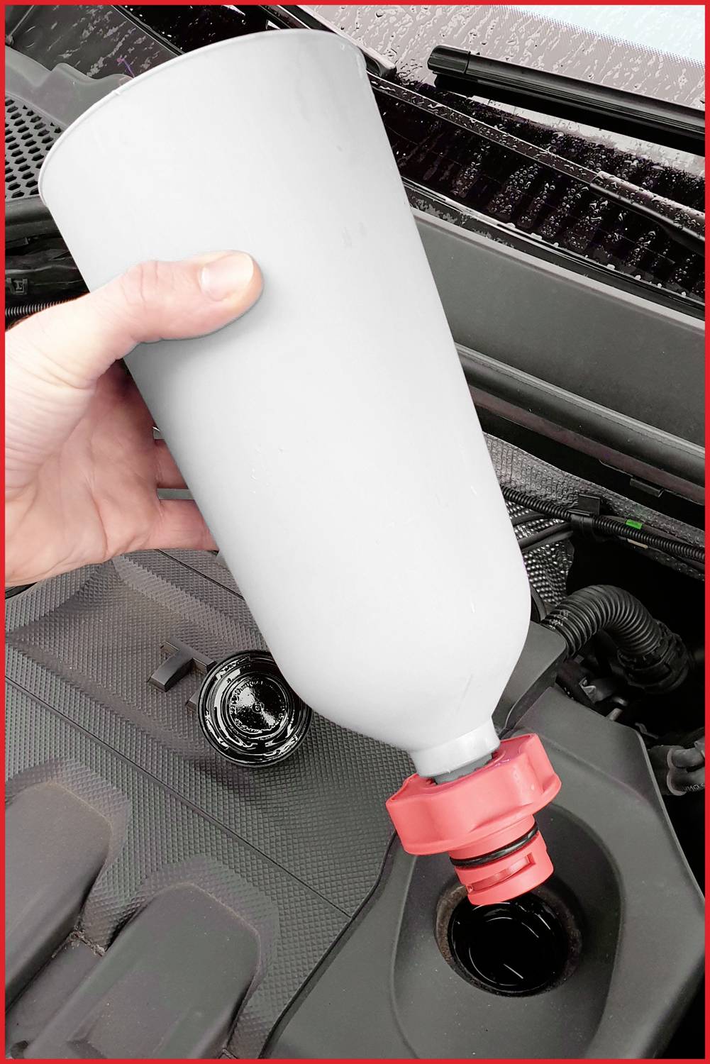 Eine Hand füllt Flüssigkeit aus einer großen grauen Flasche mit rotem Verschluss in das Einfüllloch des Fahrzeugmotors.