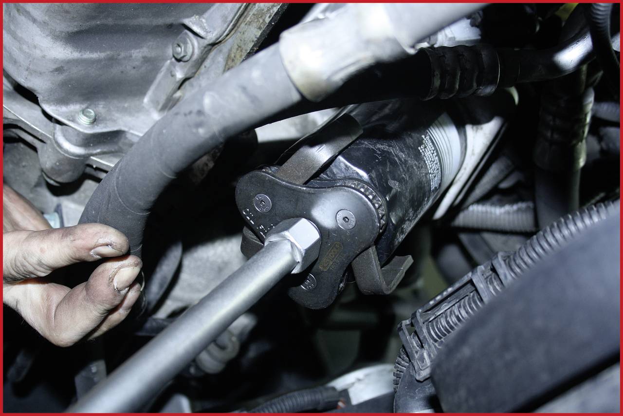Mechanikerhand benutzt Werkzeug zum Lösen eines Ölfilters im Motorraum. Umgebung zeigt Kabel und Schläuche.
