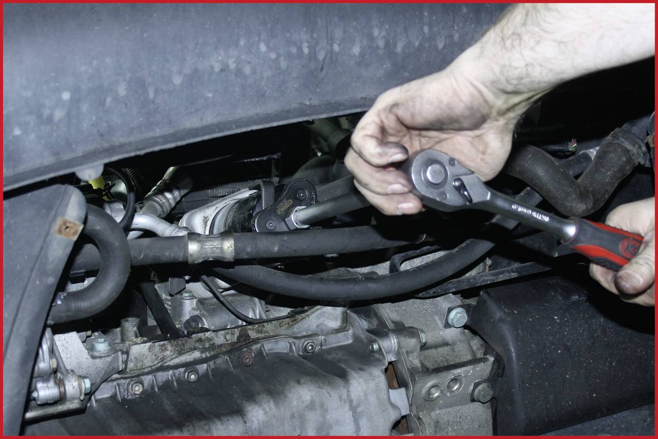 Ein Mechaniker mit Handschuhen repariert den Motor eines Fahrzeugs mit einem Schraubenschlüssel. Er arbeitet an Rohren und Kabeln.