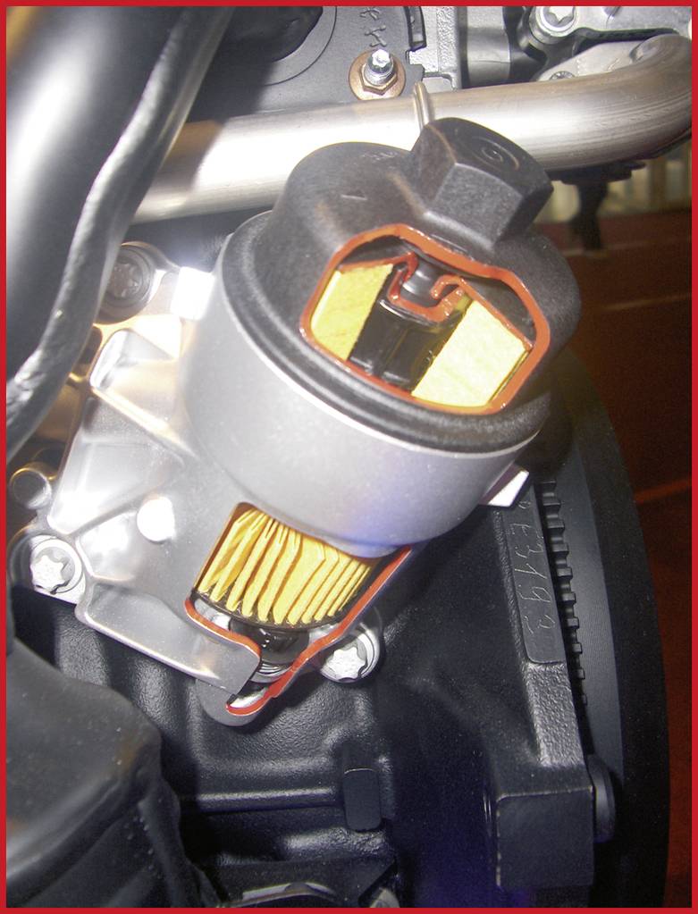 Ein silberner Ölfilter mit gelbem Filterelement ist in einer metallischen Halterung an einem schwarzen Motor angebracht.