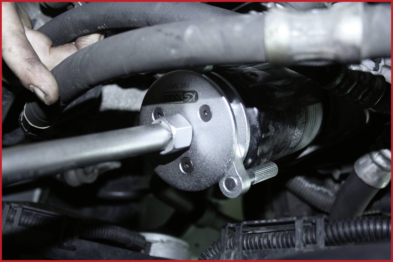 Eine Person hält eine Autoteil-Komponente mit Schläuchen und Metallbefestigung. Details von Motorteilen sind sichtbar.