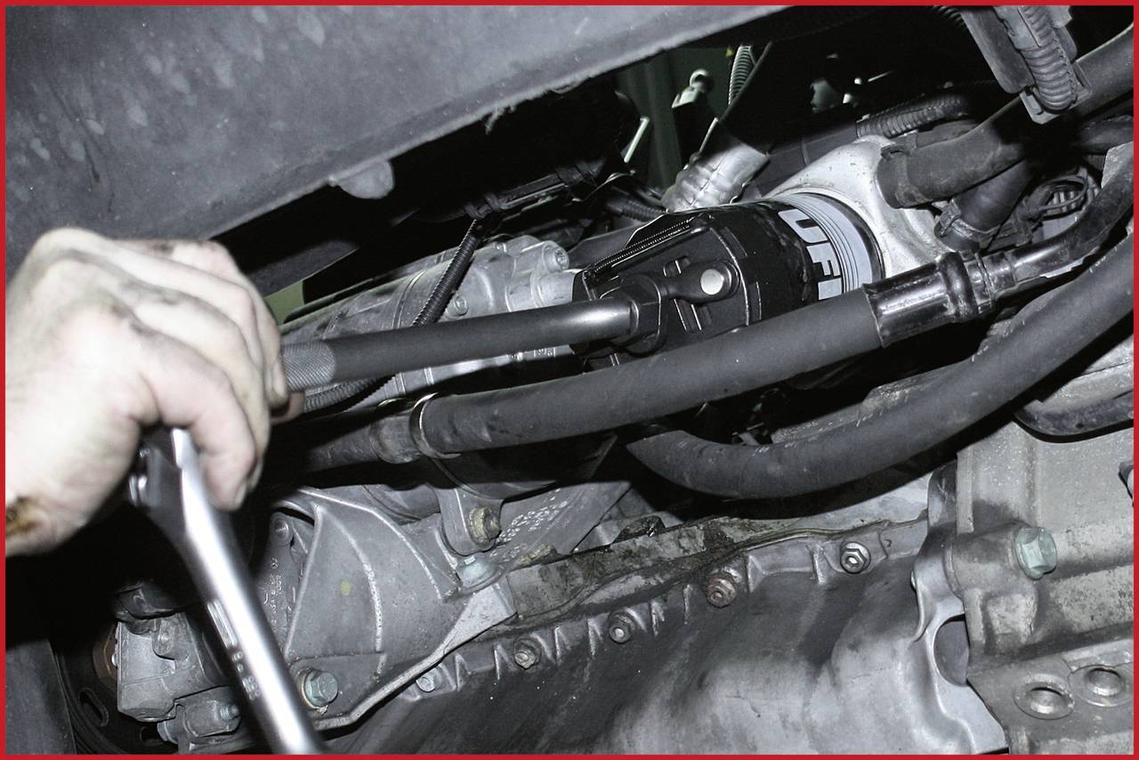 Nahaufnahme einer Hand, die mit einem Schraubenschlüssel an einem Fahrzeugmotor arbeitet, sichtbar ist ein Ölfilter.