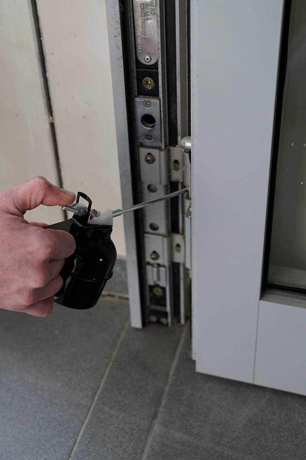 Eine Hand hält ein Werkzeug, das in einen Türrahmen eingeführt wird, um das Schloss zu öffnen. Der Boden besteht aus grauen Fliesen.