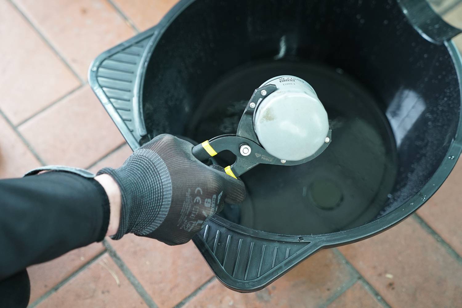 Ein schwarz gekleideter Arm mit Handschuh hält ein silbernes Gerät über einem schwarzen Plastikeimer auf einem gefliesten Boden.