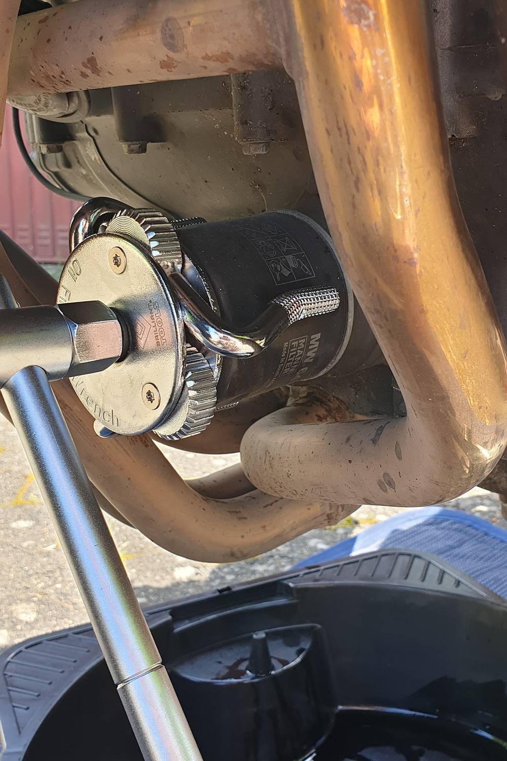 Motorölfilterwechsel: Werkzeug greift Filter zwischen Auspuffrohren, Auffangbehälter darunter.
