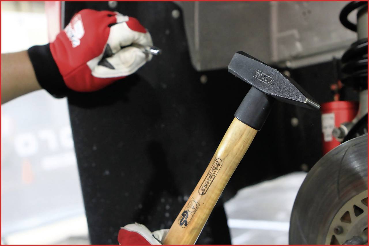 Eine Person mit roten Handschuhen hält einen kleinen Metallgegenstand, während eine andere Hand einen Hammer neben einer Bremsscheibe hält.