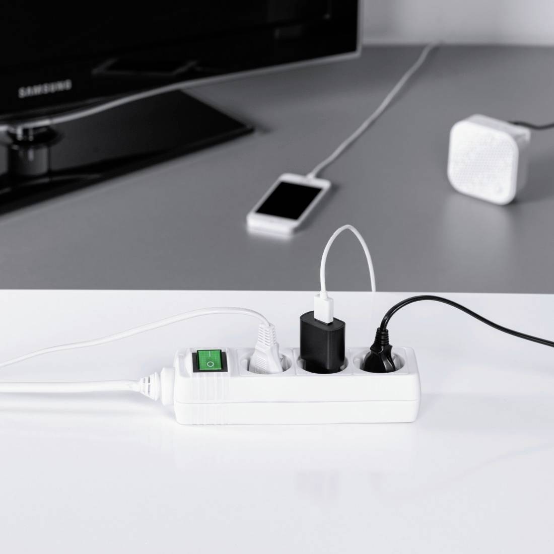 Ein weißer Mehrfachstecker mit grünem Schalter versorgt ein Smartphone-Ladegerät und zwei andere Geräte vor einem dunklen Bildschirm.