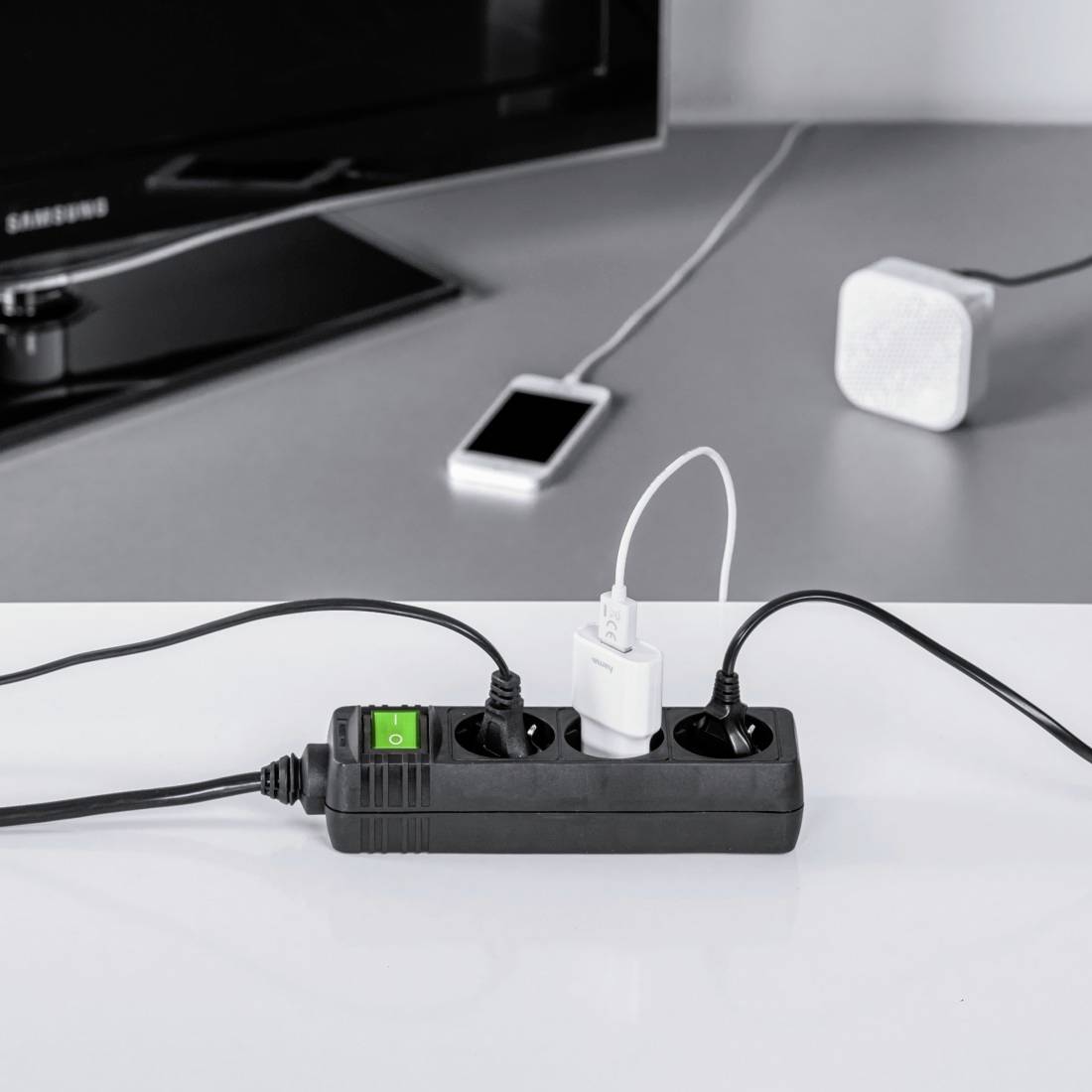 Ein Mehrfachstecker liegt auf einem Tisch, er ist mit verschiedenen Geräten verbunden, darunter ein Smartphone und ein Fernseher.