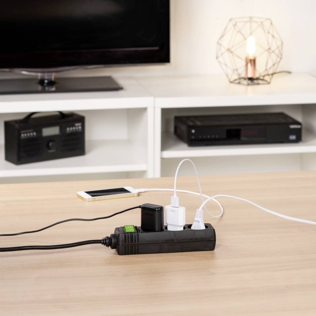 Mehrfachsteckdose auf Tisch mit vier angeschlossenen Ladegeräten für Smartphone und andere Elektronikgeräte, Lampe im Hintergrund.