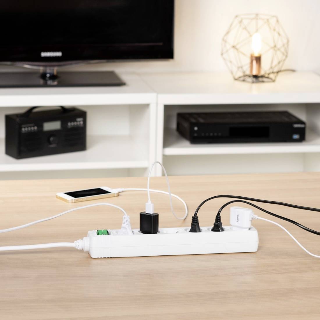 Ein mehrfach-Steckdosenleiste auf einem Tisch. Ein Smartphone lädt über ein USB-Ladegerät, umgeben von Elektronikgeräten im Hintergrund.