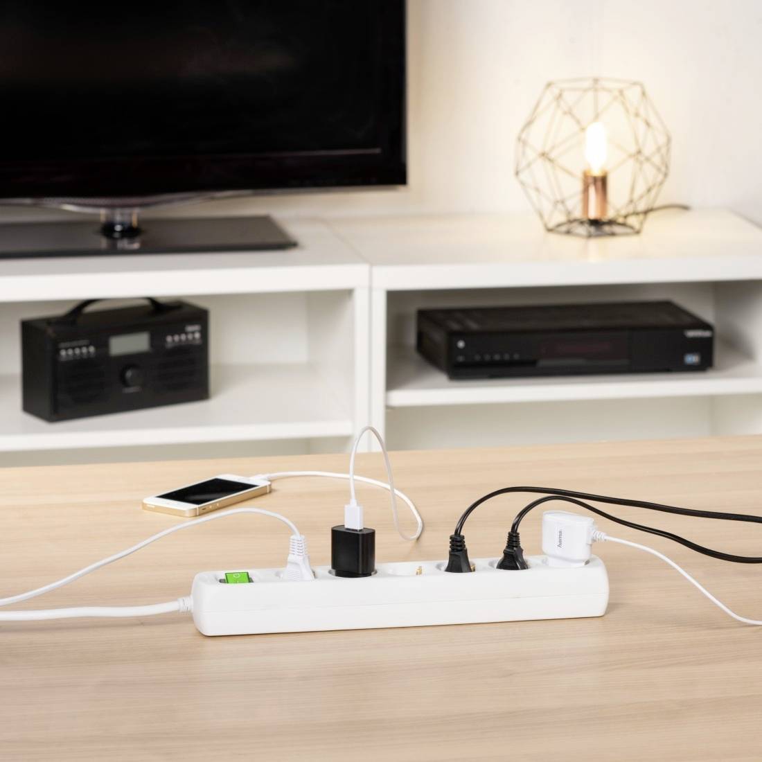 Ein Tisch mit einer Steckdosenleiste, an die mehrere Geräte angeschlossen sind. Im Hintergrund ein Fernseher und eine Lampe.